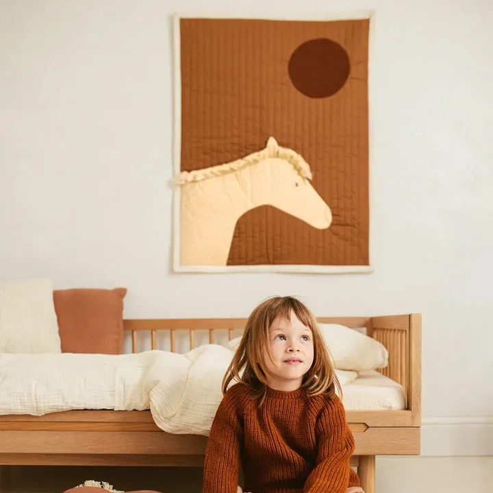 Plaid matelassé Nobodinoz avec un cheval sur fond orange, accroché au mur