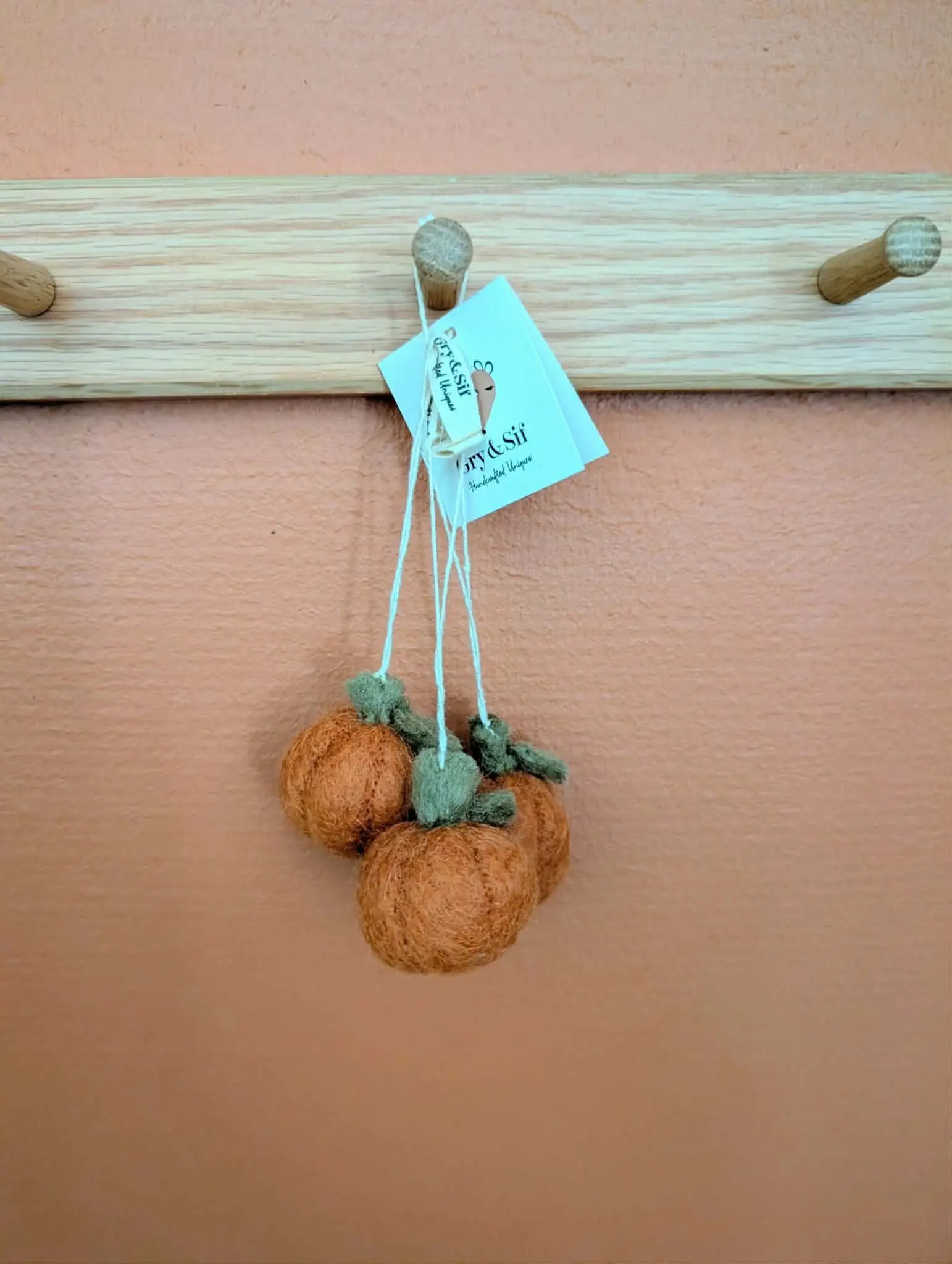Trois mini citrouilles en feutre orange Gry & Sif suspendues à une patère en bois sur un mur terracotta